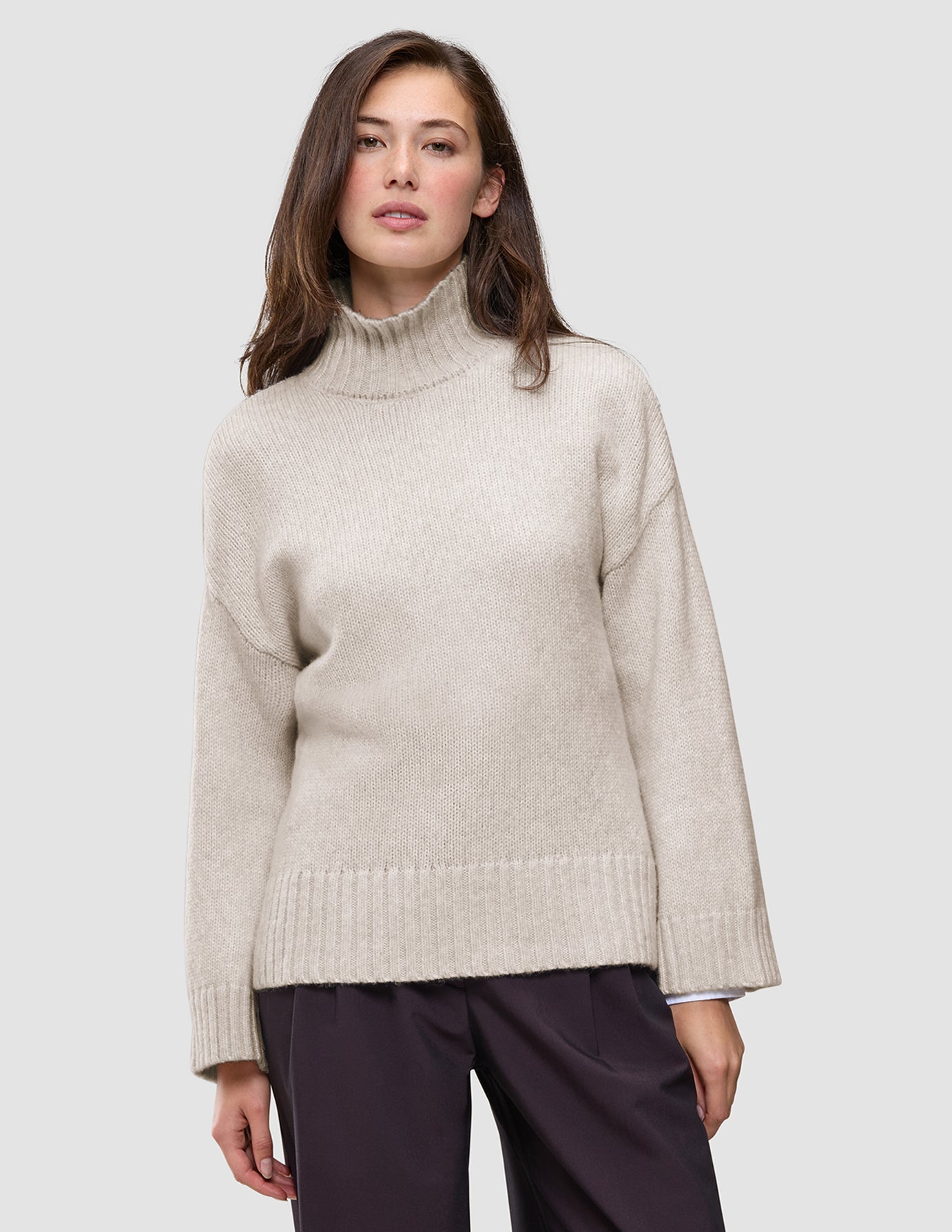 Eine Frau, die einen beigen Damen-Rollkragenpullover aus grobem, weichem Wollstrick in der Farbe Sugar Melange trägt, dazu eine dunkle Hose.
